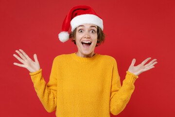 Amazed excited young Santa woman in basic yellow sweater Christmas hat keeping mouth open spreading hands isolated on red background studio portrait. Happy New Year celebration merry holiday concept.