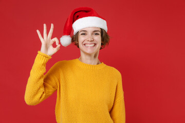 Smiling young Santa woman 20s wearing casual yellow sweater Christmas hat showing Ok gesture isolated on bright red colour background studio portrait. Happy New Year celebration merry holiday concept.