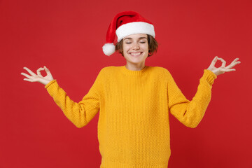 Fototapeta na wymiar Young Santa woman in yellow sweater Christmas hat hold hands in yoga gesture, relaxing meditating isolated on bright red background studio portrait. Happy New Year celebration merry holiday concept.