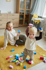 Young mother sitting on the floor and watching fo her child while he playing with toys in the room