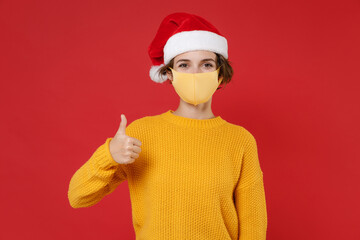 Fototapeta na wymiar Cheerful young Santa woman wearing Christmas hat face mask to safe from coronavirus virus covid-19 showing thumb up isolated on red background studio. Happy New Year celebration merry holiday concept.