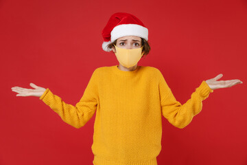 Fototapeta na wymiar Confused puzzled young Santa woman in Christmas hat face mask safe from coronavirus virus covid-19 spreading hands isolated on red background studio. Happy New Year celebration merry holiday concept.