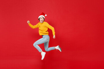 Full length side view of funny young Santa woman in yellow sweater Christmas hat jumping like running looking aside isolated on red background studio. Happy New Year celebration merry holiday concept.