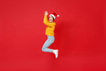Fototapeta na wymiar Full length side view of excited young Santa woman in yellow sweater, Christmas hat jumping rising hands isolated on red background studio portrait. Happy New Year celebration merry holiday concept.