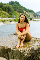 Fototapeta na wymiar A young brunette girl in a red swimsuit sits on the stones by the sea.