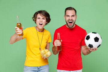 Screaming angry young couple friends sport family woman man football fans in t-shirts cheer up support favorite team with soccer ball hold beer bottles bowl chips isolated on green background studio.