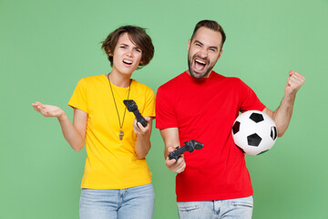 Displeased happy couple friends sport family woman man football fans in t-shirts cheer up support favorite team with soccer ball play pc game with joystick console isolated on green background studio.
