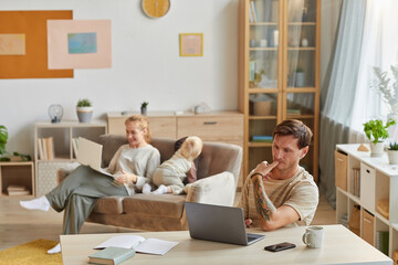 Serious man sitting at the table and concentrating on his online work on laptop with his family in the background at home