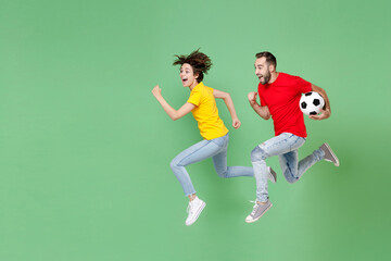 Fototapeta na wymiar Full length of excited young couple friends sport family woman man football fans in t-shirts cheer up support favorite team with soccer ball jumping like running isolated on green background studio.
