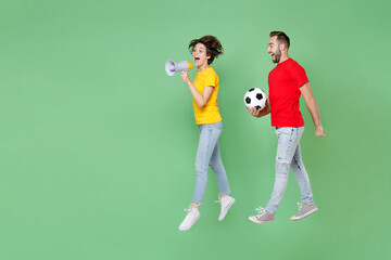 Fototapeta na wymiar Full length of excited couple friends sport family woman man football fans in t-shirts cheer up support favorite team with soccer ball jumping scream in megaphone isolated on green background studio.