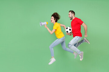 Fototapeta na wymiar Full length of cheerful couple friends sport family woman man football fans in t-shirts cheer up support favorite team with soccer ball jumping scream in megaphone isolated on green background studio.