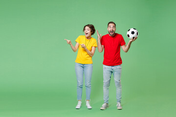 Full length surprised couple friends sport family woman man football fans in yellow red t-shirts cheer up support favorite team with soccer ball point index fingers aside isolated on green background.