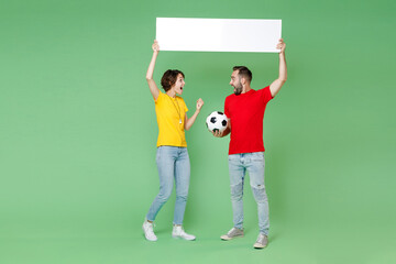 Full length shocked couple friends sport family woman man football fans in t-shirts cheer up support favorite team with soccer ball hold big white empty blank billboard isolated on green background.