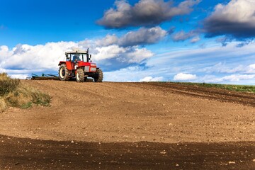 Obraz premium Red tractor cultivates the soil in the field with a cultivator after harvest. Autumn sunny day. Agricultural landscape in the Czech Republic. Work on the farm.