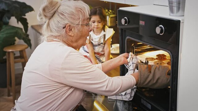 Granny is opening the oven and taking out hot cookies, surprised grandchild is looking at it. Baking homemade biscuits at modern kitchen