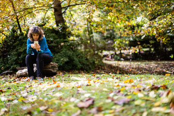 Autumn scene of middle aged woman wearing blue sweater sitting on stone and looking at smartphone next to pond covered in fallen leaves