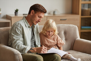 Father sitting on sofa with his little son and teaching him to draw in the room