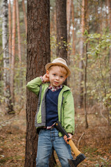 Little boy in a hat in the autumn forest. A boy with an ax near a tree. Woodcutter hold ax. Standing in the forest with axe.