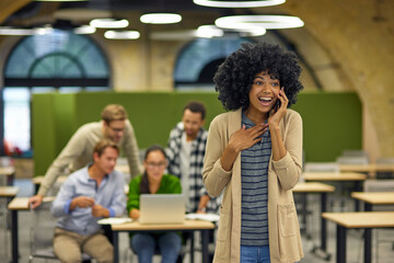 Exciting news. Young overjoyed mixed race woman talking on smartphone and smiling while standing in the coworking space with colleagues working on the background