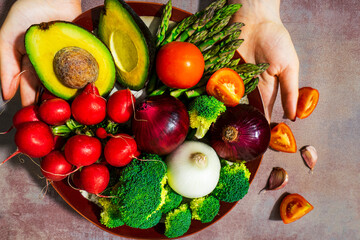Plate with organic and local vegetables freshly picked from the garden served by women's hands. Composition with broccolis, asparagus, radish, onions, red tomatoes, garlic, avocado on light background