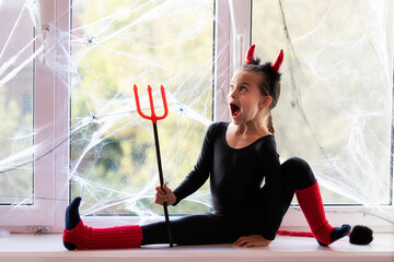 Little girl gymnast in costume of little devil playing about and stretching on window sill