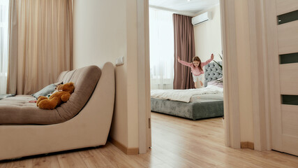 A cute girl jumping down on a big bed in a bedroom having her teddybear in a guestroom