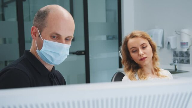 Professional male dentist examining young woman and showing treatment dental issues on computer screen x-ray image in stomatology clinic indoors. Healthcare. Medicine.