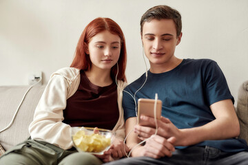 Couple of teenagers sitting on the couch at home, watching something on the phone, using the same pair of earphones while spending time together