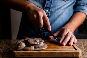 Man preparing culinary seafood, cutting raw shrimp on a wood table with a knife.