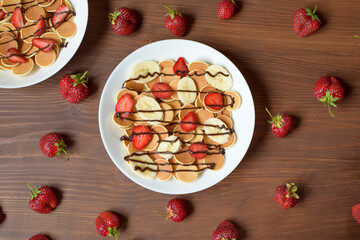 Mini pancakes with strawberries and chocolate lie on a wooden table.