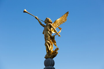 Side view of golden statue of angel blowing in trumpet seen against bright blue sky,...