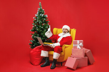 Excited Santa Claus man in Christmas at suit sit in armchair with fir tree presents work on laptop pc computer showing thumb up isolated on red background. Happy New Year celebration holiday concept.
