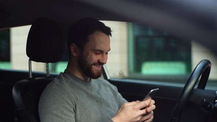 Tracking left shot of cheerful young man sitting in car and typing online message on cellphone, side view. Smiling businessman using phone in auto. Male sitting in vehicle and works on smartphone.