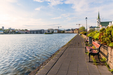 Footpath lined with empty wooden benches along the shore of a lake in a city centre. Reykjavik,...
