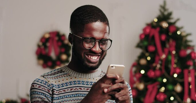 Portrait of happy young african american man in glasses and winter sweater holding phone in hands and typing message on christmas tree background.