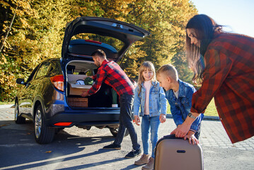 Joyful lovely young family loading the auto's trunk by carton boxes and suitcases while relocating...
