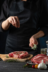 Close-up view of chef pours rosemary on raw steak on wooden chopped board. Backstage of preparing grilled pork meat at restaurant kitchen on dark blue background. Frozen motion. Vertical format.