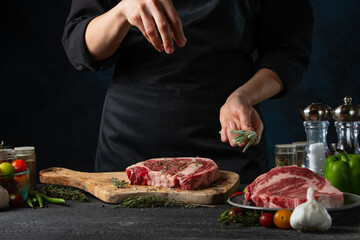 Obraz na płótnie Canvas Close-up view of chef pours rosemary on raw steak on wooden chopped board. Backstage of preparing grilled pork meat at restaurant kitchen on dark blue background. Frozen motion. Horizontal format.