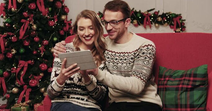 Portrait of happy young family looking at photo on tablet sitting at home on sofa near Christmas tree.