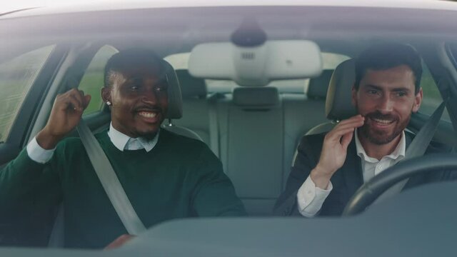 Two happy young attractive businessmen listening music dancing in car at sunlight. Feel happy. Talking about business. Wheel automobile transport. Slow motion