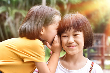 Two little sister girls whisper in ear at park outdoor.