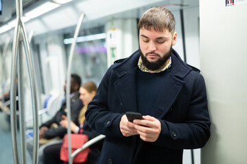 Young focused man browsing and typing messages on phone on way to work in modern metro car