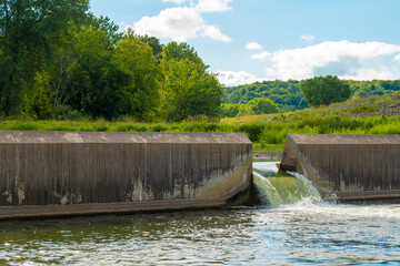 Water flows through the floodgate in a concrete dam on a river, with green trees and vegetation on the hills in the background. © scandamerican