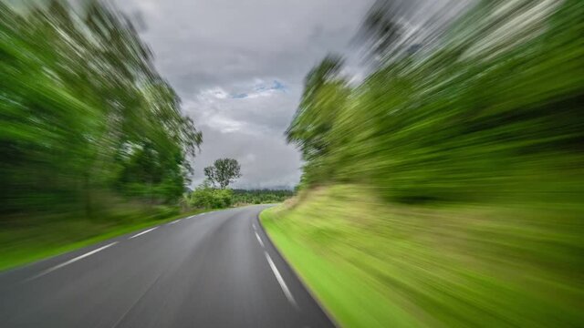 A drive on the narrow road in Norway. The road is running through the forest and meadows. The vegetation is lush and green. Asphalt is wet after the rain. Gray clouds hanging above.