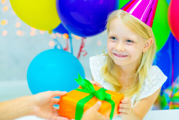A girl in a festive cap against a background of colorful balloons with a smile takes a gift from...