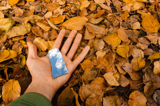 Holding Broken Mirror Splinter On Hand Palm Which Reflects Blue Sky With White Clouds On Bright Fall Leaves Background. Copy Space. Top View