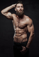 Fototapeta na wymiar Brutal bearded guy with naked torso and muscular build with hand in pocket and hand under head posing in dark background.