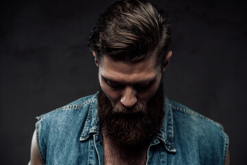 Dressed in modern casual clothing brutal bearded guy with stylish haircut and brown hairs posing looking down in dark background.