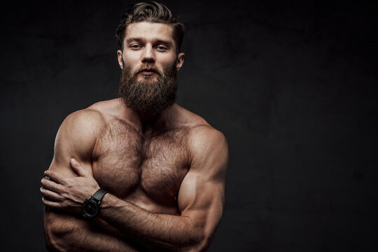 Naked man with muscular build and beard posing with crossed arms looking at camera in dark background.
