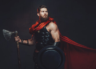 Warlike and armoured rome empire fighter with beard and muscular build posing holding shield and axe in dark room with spotlight.